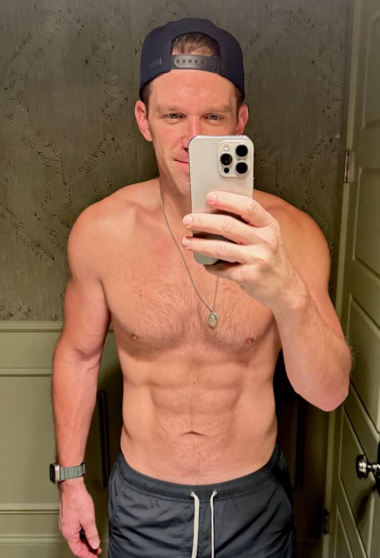 A shirtless man, possibly an online fitness coach, wearing a backward cap and necklace, takes a mirror selfie in a bathroom while holding his smartphone and wearing dark shorts.